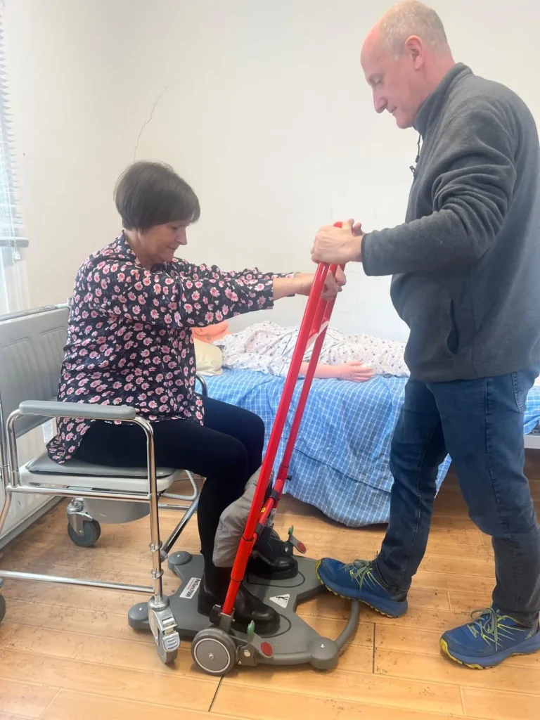 A carer helping a client to stand using a mobility hoist beside her bed